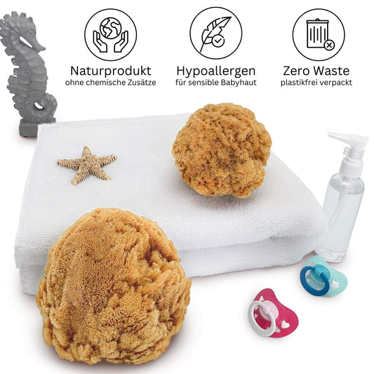 Zwei naturbelassene Babyschwämme aus dem Mittelmeer auf weißem Handtuch, umgeben von Schnuller, Seifenspender und Seestern – ideal für sensible Babyhaut