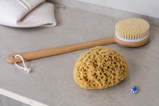 Eine langstielige Bürste liegt neben einem Honeycomb-Naturschwamm auf einer hellen Ablage.