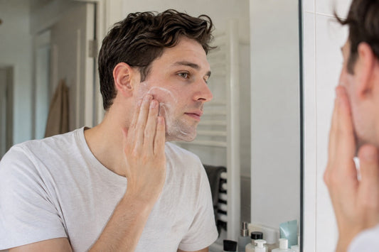 Ein Mann verteilt vor dem Badspiegel schaumigen Gesichtsreiniger mit den Fingerkuppen auf der Wange – sanft reinigen, statt gegen Pickel zu schrubben.