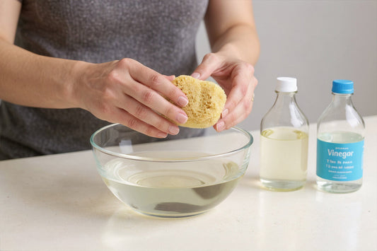 Menstruationsschwamm reinigen: Zwei Hände drücken einen Menstruationsschwamm über einer Glasschale aus, daneben eine Flasche mit heller Flüssigkeit und eine Essigflasche