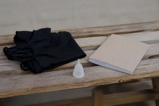 Gefaltete schwarze Bademode, eine Menstruationstasse und ein Heft liegen auf einer Holzbank.