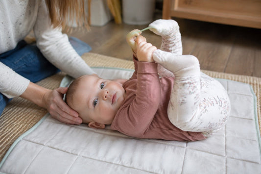 Trockene Babyhaut: Ein waches Baby in rosa Oberteil liegt auf einer gesteppten Unterlage, hält ein kleines Spielzeug und greift nach seinem Fuß, eine Frau legt ihm die Hand an den Kopf
