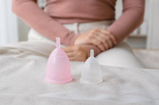 Eine rosa und eine kleinere transparente Menstruationstasse stehen auf hellem Stoff.