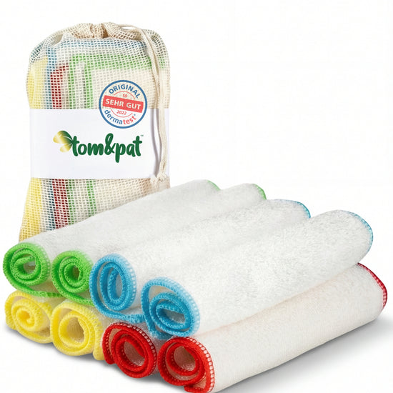 Acht gerollte Bambus-Putztücher mit grüner, gelber, blauer und roter Kante neben einem Baumwollbeutel mit tom&pat-Banderole
