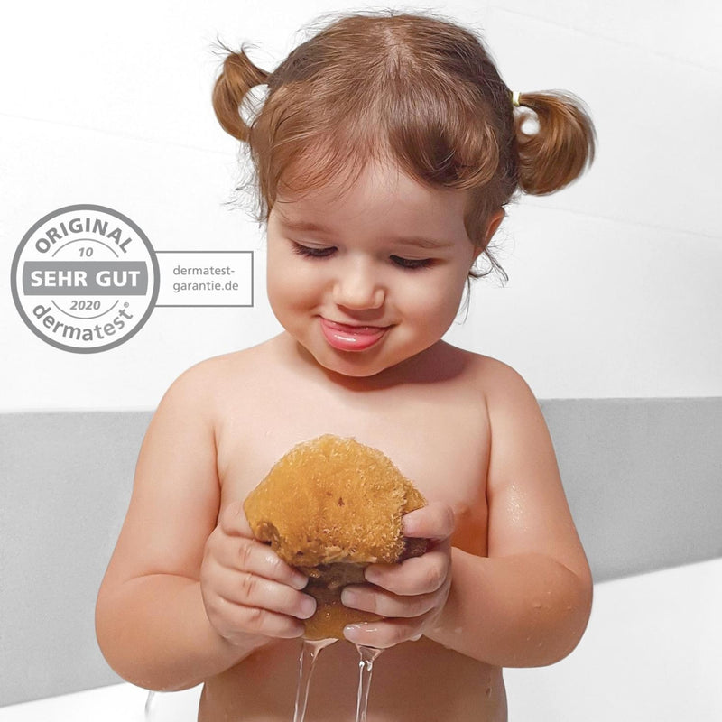 Kleinkind mit Zöpfen in der Badewanne drückt einen tropfnassen Babyschwamm aus Naturschwamm, Dermatest-Siegel sehr gut