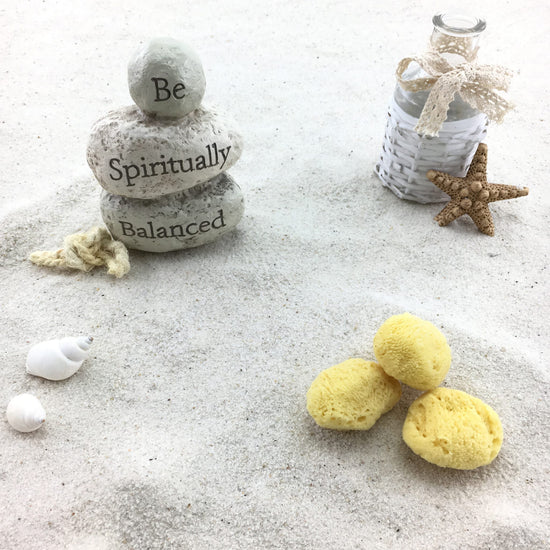 Drei gelbe Kosmetikschwämme im hellen Sand neben Steinturm mit Aufschrift "Be Spiritually Balanced", Muscheln und Seestern