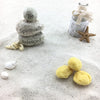 Drei gelbe Kosmetikschwämme im hellen Sand neben Steinturm mit Aufschrift "Be Spiritually Balanced", Muscheln und Seestern