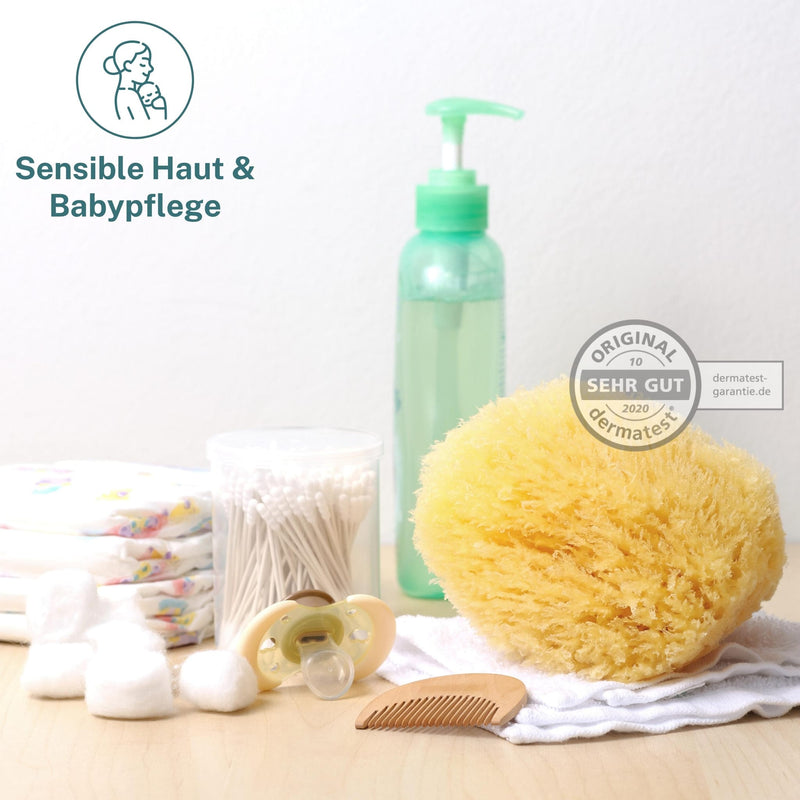 Naturschwamm Grass zwischen Babypflege-Sachen wie Windeln, Wattestäbchen, Schnuller und Holzkamm, daneben das Dermatest-Siegel „sehr gut“